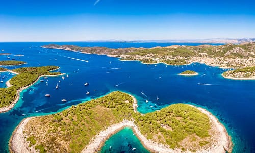 Aerial view of Hvar Islands in Croatia, showcasing turquoise waters, lush greenery, and numerous sailboats under a clear blue sky.