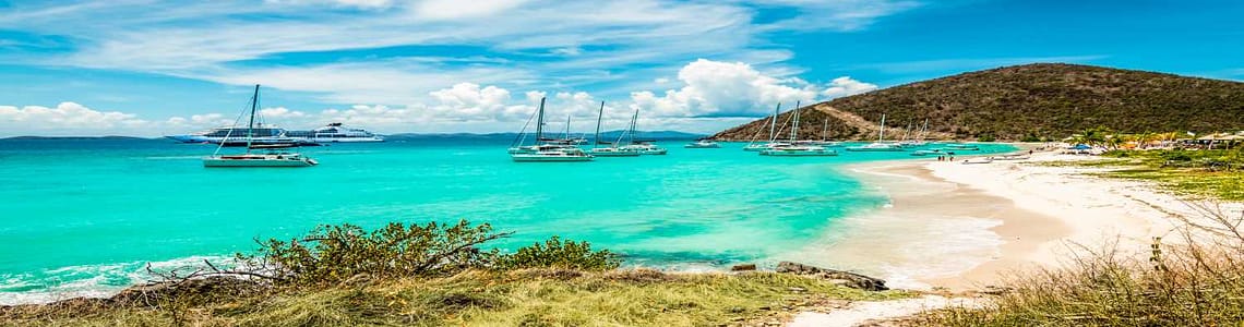 Tropical beach with turquoise water, sailboats anchored near the shore, and a hillside landscape under a blue sky.