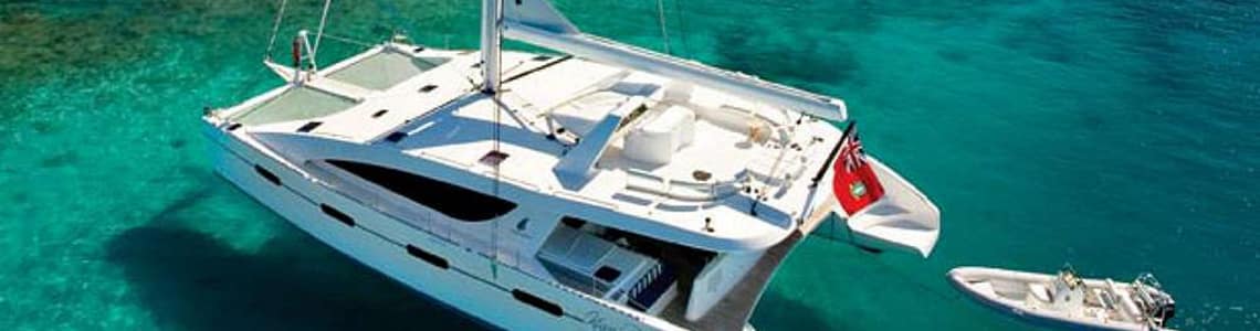 Luxury catamaran anchored in crystal-clear turquoise waters with a small dinghy attached, near a sandy beach.