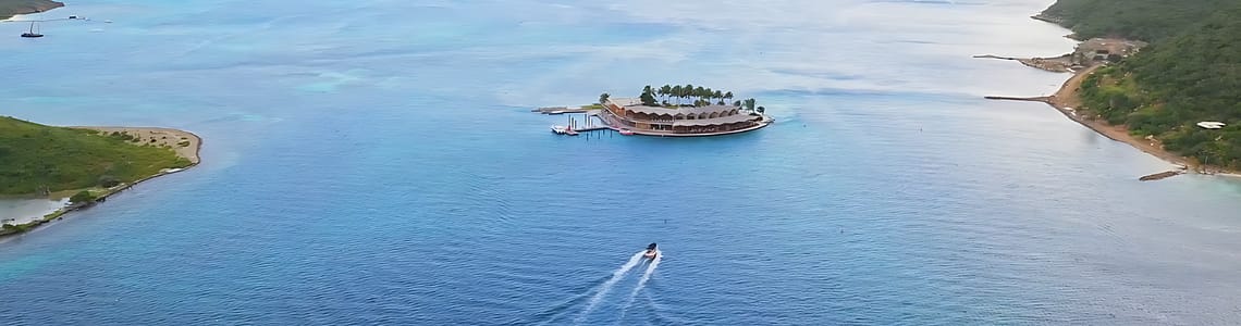 Aerial view of an island resort surrounded by turquoise ocean waters, featuring lush greenery and a boat approaching the dock.
