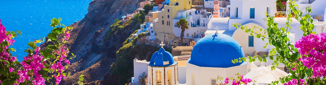 Scenic view of Santorini, Greece with iconic blue-domed churches, vibrant white buildings, and blooming pink flowers overlooking the Aegean Sea.