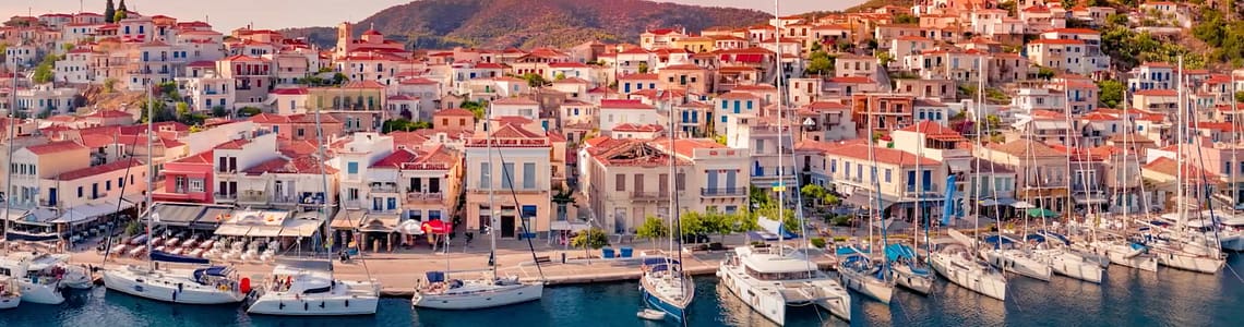 Scenic view of a picturesque coastal village with colorful houses and sailboats docked in the harbor at sunset, surrounded by lush hills.