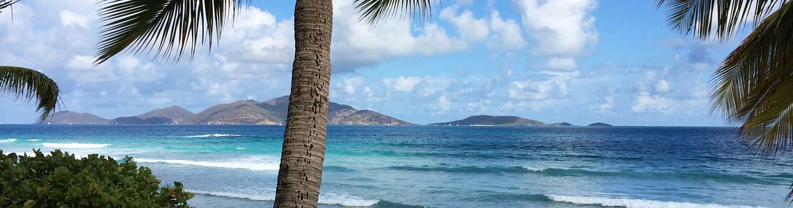 Tropical beach with palm trees, clear blue sea, and distant islands under a sunny sky.