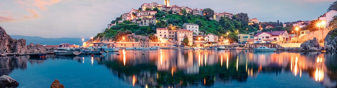 Scenic view of a charming hillside village at sunset with colorful sky, illuminated buildings, and reflective water in a tranquil harbor.