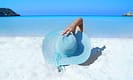 Person relaxing on a sandy beach near clear blue sea with a large sun hat.