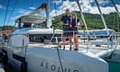 Couple standing on sailing yacht Aeolus at marina with green hillside and blue sky background.