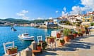 Scenic view of a charming Greek coastal village with colorful houses, boats in the turquoise sea, and outdoor seating adorned with potted flowers under a clear blue sky.