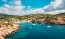 Scenic view of a coastal bay with turquoise waters, rocky cliffs, and sailboats anchored near a Mediterranean island, under a partly cloudy sky.