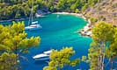 Scenic view of a tranquil bay with turquoise waters and anchored sailboats surrounded by lush greenery and rocky hills in a Mediterranean landscape.