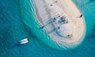 Aerial view of a small sandy island with people gathered and a boat anchored nearby in clear turquoise water, ideal tropical destination.