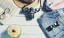 Travel essentials including sunglasses, camera, straw hat, open book, denim shorts, scarf, and a fresh coconut drink on a white wooden table.