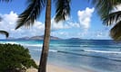 Tropical beach with palm trees, clear blue sea, and distant islands under a sunny sky.