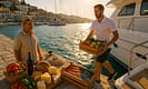 Crew member loads fresh Greek produce, wine, bread, cheese, and seafood onto a yacht at a sunlit Greek marina with a local supplier, dock rope, and the sparkling Aegean Sea in the background.