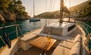 Modern white yacht anchored in a turquoise Greek cove at golden hour, with sunlit teak deck, wine bottle, and village on hillside. Pine trees, limestone cliffs, and serene waves highlight the unforgettable, luxurious yacht holiday experience in Greece.
