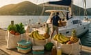 Provisioning for a BVI yacht vacation: reusable bags filled with fresh tropical produce, food containers, and bottled drinks on a weathered dock table, with a crew member loading supplies onto a sailboat under golden sunlight and Tortola hills in the background.
