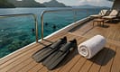 Luxury yacht teak deck with dive fins, white towel, and sun loungers overlooking turquoise Indonesian waters, visible coral reefs, scuba divers below, and lush islands in the background under golden sunlight.