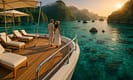 A luxurious private yacht anchored in clear turquoise waters of Raja Ampat, Indonesia, with a couple enjoying the sunset. Pristine decks, lush islets, and limestone cliffs surround the scene, while coral and tropical fish are visible beneath the water’s surface.