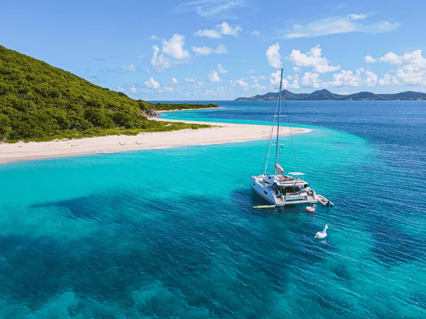 Luxury catamaran anchored near a tropical white sand beach with turquoise water and lush green hills in the Caribbean under a clear blue sky.