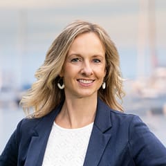 Professional woman smiling outdoors with blurred boats in background, wearing a blue blazer and white top, showcasing a confident and approachable demeanor.