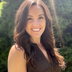Smiling woman with long brown hair outdoors in sunlight, standing in front of green foliage.