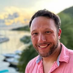 Smiling man in pink shirt by a scenic waterfront at sunset, with blurred greenery and boats in the background.
