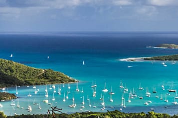 View of a scenic Caribbean bay with numerous sailboats on turquoise waters, surrounded by lush green islands under a partly cloudy sky.