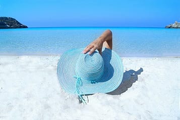 Person relaxing on a sandy beach near clear blue sea with a large sun hat.