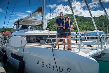 Couple standing on sailing yacht Aeolus at marina with green hillside and blue sky background.