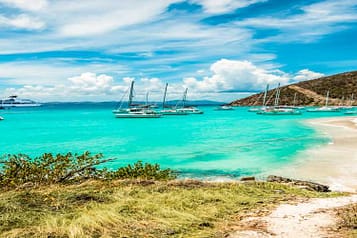 Tropical beach with turquoise water, sailboats anchored near the shore, and a hillside landscape under a blue sky.