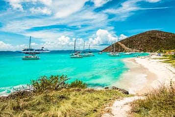 Tropical beach with turquoise waters, sailboats, and a hilly landscape in the British Virgin Islands.