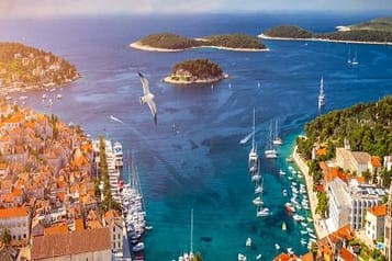 Scenic aerial view of Hvar, Croatia, featuring vibrant orange rooftops, clear blue Adriatic Sea, sailboats, yachts, and lush green islands.