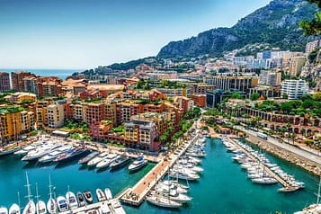 Aerial view of Monaco harbor with luxury yachts, waterfront buildings, and surrounding hills.