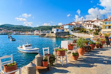 Scenic view of a charming Greek coastal village with colorful houses, boats in the turquoise sea, and outdoor seating adorned with potted flowers under a clear blue sky.