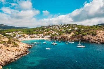 Scenic view of a coastal bay with turquoise waters, rocky cliffs, and sailboats anchored near a Mediterranean island, under a partly cloudy sky.