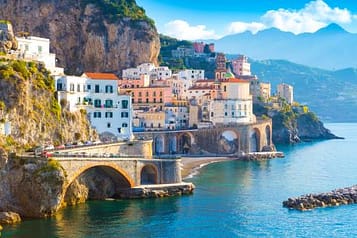 Scenic view of Amalfi Coast, Italy, featuring colorful cliffside buildings, arched bridge, and tranquil blue sea with mountainous backdrop.