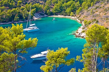 Scenic view of a tranquil bay with turquoise waters and anchored sailboats surrounded by lush greenery and rocky hills in a Mediterranean landscape.