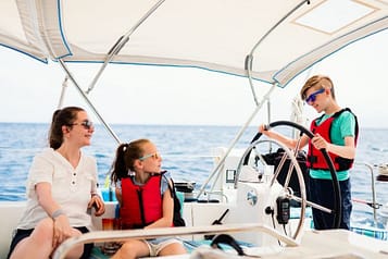 Family enjoying sailing on a sunny day, children wearing life vests and sunglasses, open sea background.