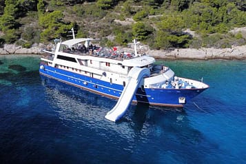 Luxury yacht with waterslide anchored near a scenic coastline with clear turquoise water and lush green trees.