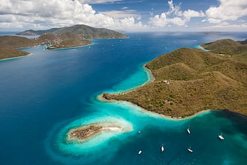 Aerial view of tropical islands with lush green hills and turquoise waters, surrounded by boats and expansive ocean, under a partly cloudy sky.