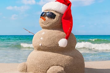 Sand snowman wearing sunglasses and a Santa hat on a sunny beach with ocean waves in the background.