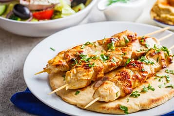 Grilled chicken skewers garnished with herbs on pita bread, served with a Greek salad and hummus on a rustic table setting.
