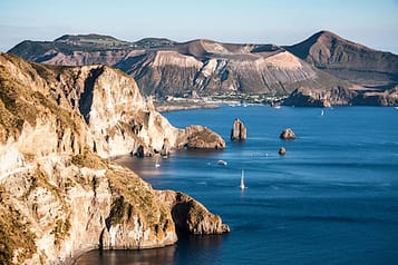 Scenic view of rocky coastline and calm sea with sailboats near the Aeolian Islands, Italy, showcasing natural beauty and serene landscape.
