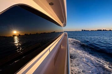 Luxury yacht cruising at sunset with city skyline and ocean waves, reflecting the vibrant evening sky.