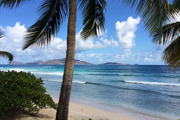Tropical beach with palm trees, clear blue sea, and distant islands under a sunny sky.