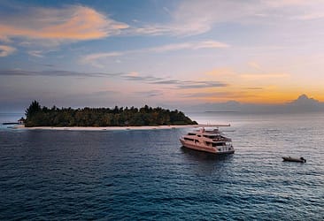 Luxury yacht cruising at sunset near a tropical island with lush greenery and clear blue water.