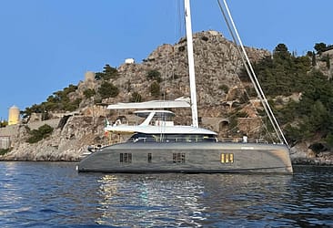 Luxury yacht anchored near rocky coastline with hillside windmills, clear sky, and calm water.