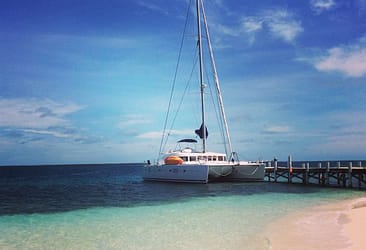 Sailboat docked by sandy beach with clear turquoise water and blue sky background.