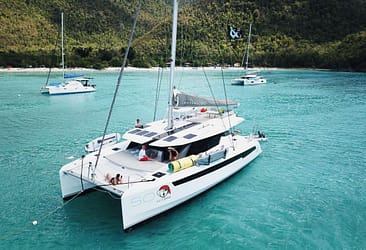 Luxury catamaran anchored in turquoise waters near lush green mountains, featuring people relaxing on deck.