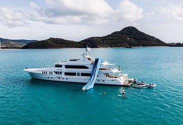 Luxury yacht with a water slide anchored in turquoise waters near a scenic island, featuring people paddleboarding and relaxing on a sunny day.