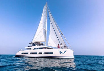Luxury sailing catamaran on open sea with three passengers, bright blue sky, and full sails.